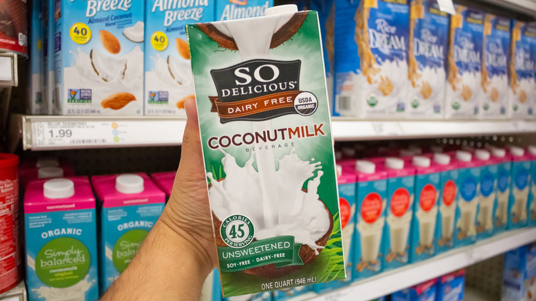 A hand holds up a box of So Delicious coconut milk in front of a grocery store shelf of plant-based milks