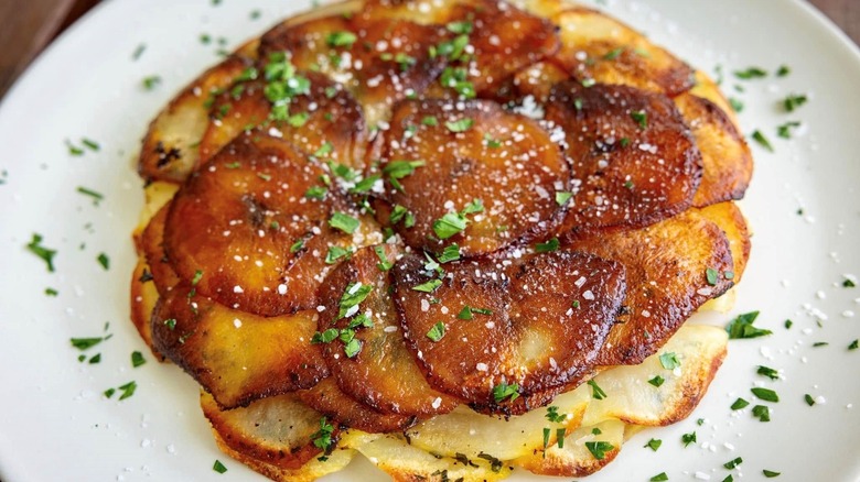 Browned layered round potato slices in a round design (Pommes Anna) sprinkled with parsley and salt on a round white dish