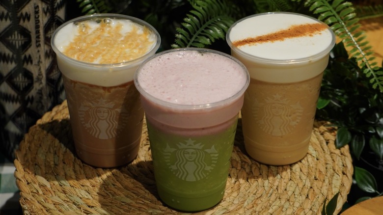 Overhead shot of Starbucks' Salted Caramel Mocha Strato Frappuccino, Strawberry Matcha Strato Frappuccino, and Brown Sugar Strato Frappuccino placed on a woven placemat in front of greenery