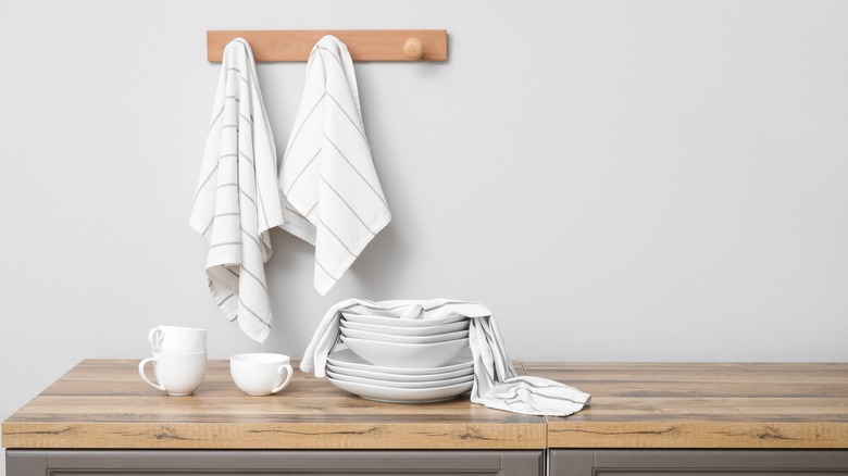 White dish towels hanging on a wooden board above white dishware with another towel draped over