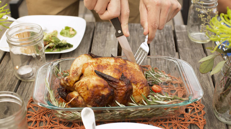 A person cuts into a full grilled chicken with a knife and fork at a wooden outdoor table