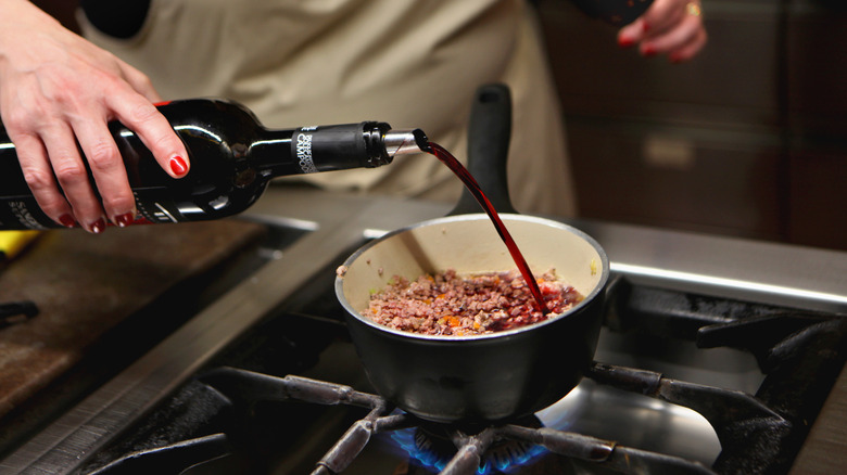 Hand pouring red wine into a pot of ground meat cooking on the stove