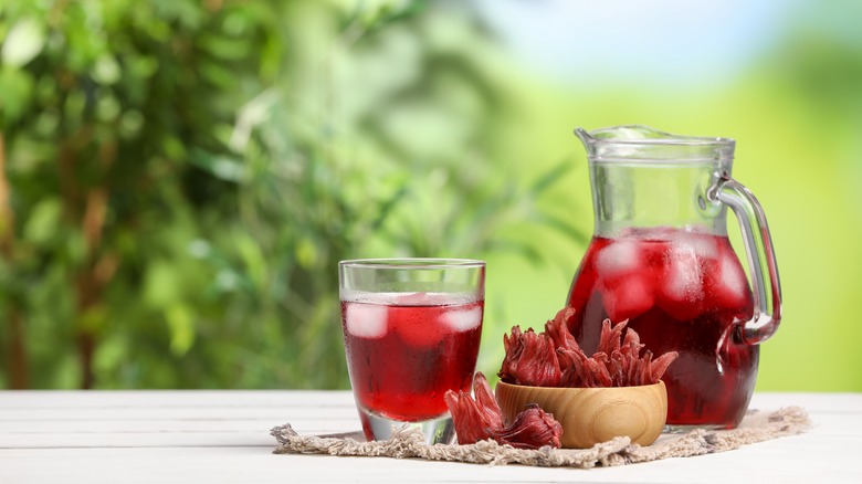 A glass and pitcher of hibiscus iced tea next to a bowl of dried hibiscus flowers