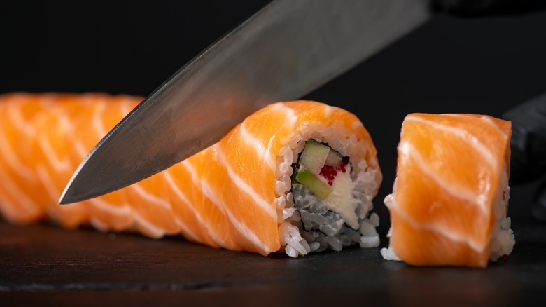Knife cutting sushi roll wrapped with raw salmon
