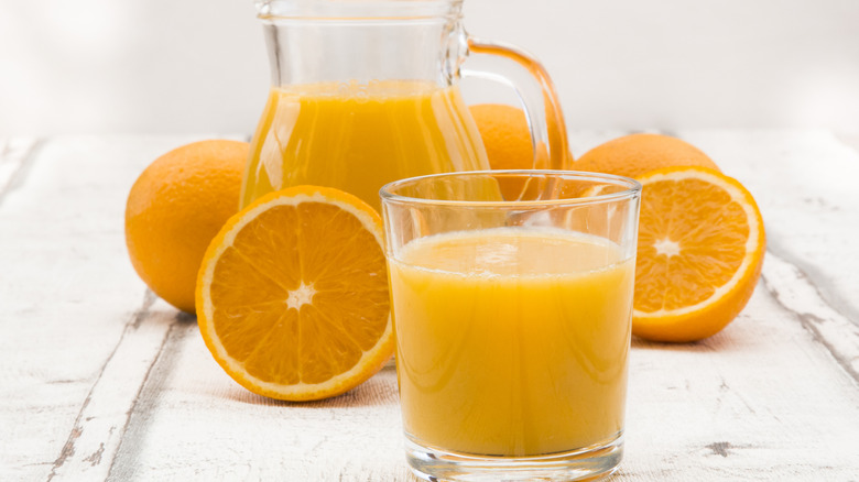 A glass of orange juice sits in the foreground, behind it sits oranges and a pitcher of OJ