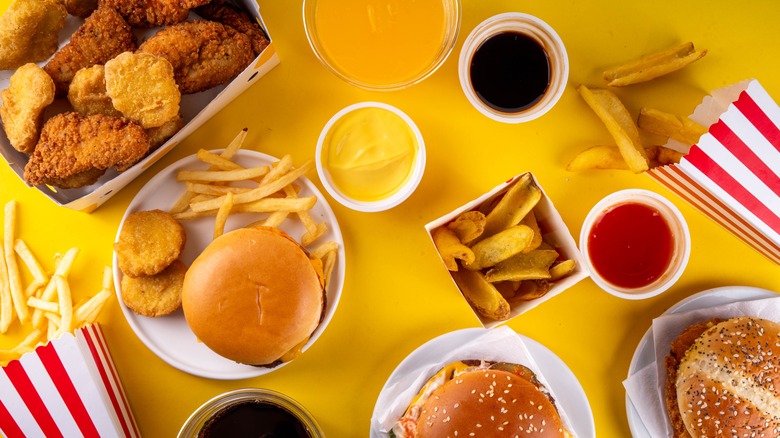 A collection of fast food burgers, fries, and chicken nuggets on plate surrounded by dipping sauces on a yellow table