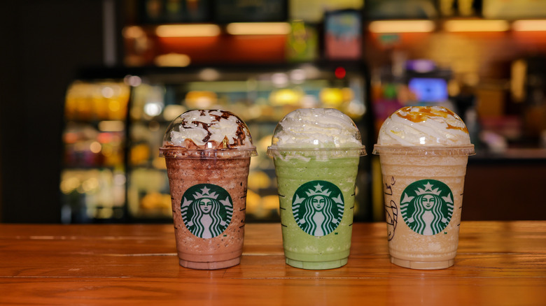 Three different Starbucks Frappuccinos on a wooden table with a blurred Starbucks store interior in the background.