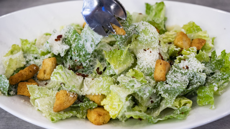 Tongs set down more lettuce onto a plate of Caesar salad