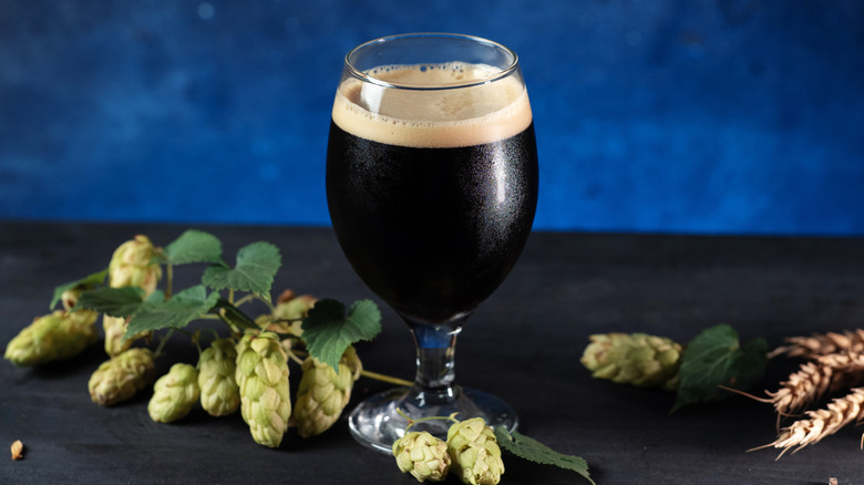 Large glass of dark beer on a dark surface with hops on one side