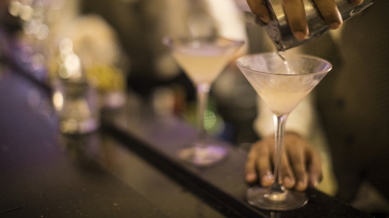 hands pouring a dirty martini from a shaker into a chilled glass on a bar