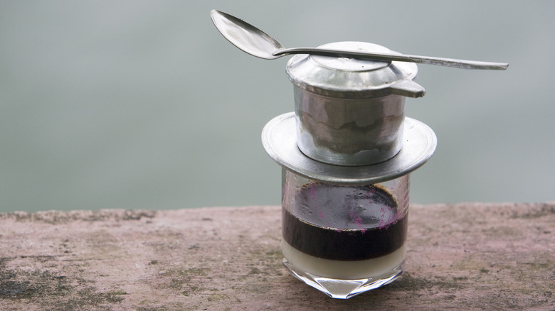 Small cup of coffee with condensed milk at the bottom, and a small metal filter known as a phin filter is on top of the glass, with a spoon on top.