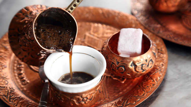 Turkish coffee being poured out of a small copper pot, into a white cup. Copper trays are holding the items, with a small bowl of sugar to the right, and a small spoon to the left. All on a gray table.