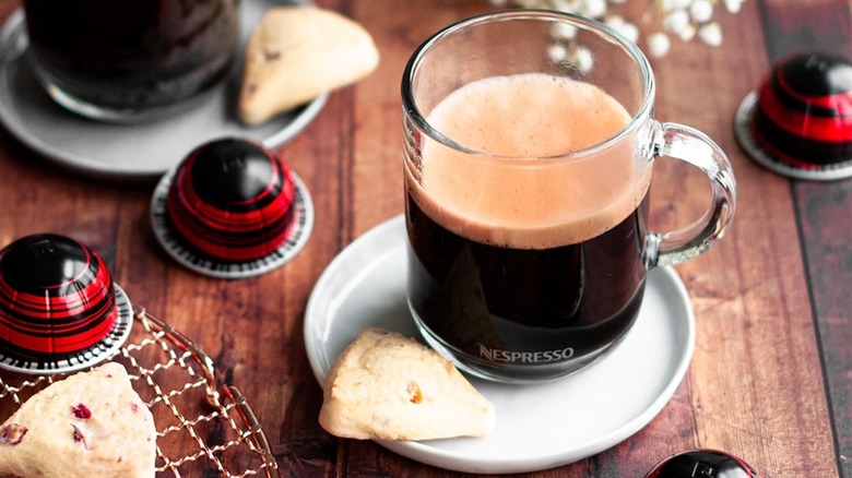 A clear mug filled with Nespresso Half Caffeinato coffee, with coffee capsules surrounding and miniature scones on a wooden table