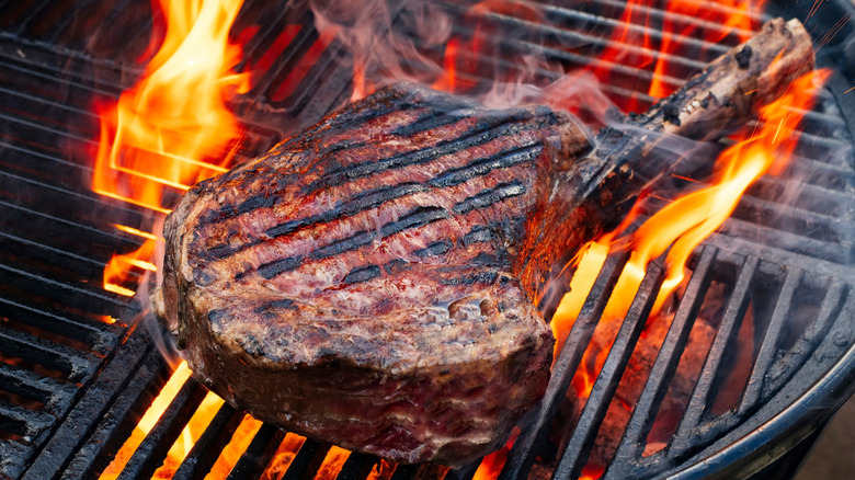 A charred tomahawk steak sits on the grill surrounded by flames