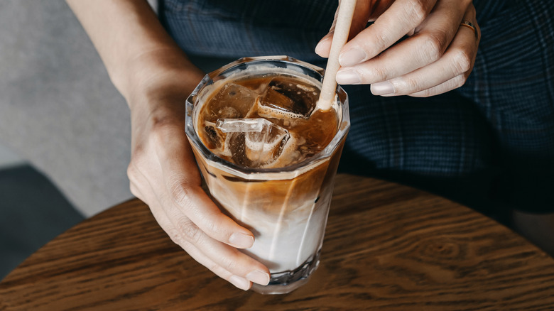 Hands stirring an iced coffee with a spoon above a wooden table
