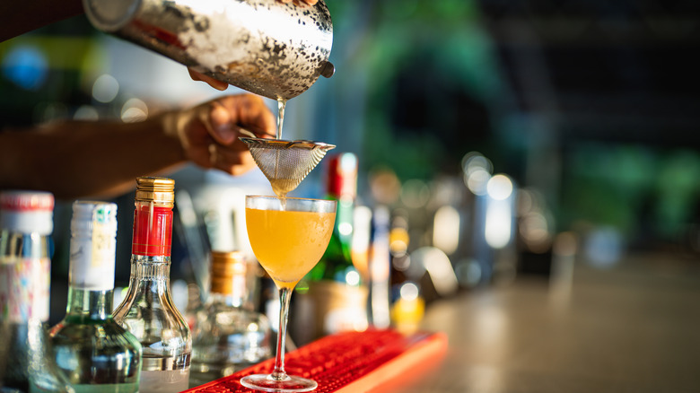 A bartender pours a drink through a strainer into a cold glass on a bar top