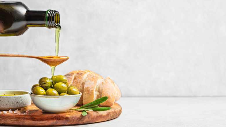 A bottle of olive oil pouring over a spoon onto a bowl of olives on a cutting board next to sliced bread