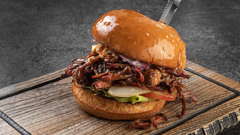 A pulled pork sandwich on a cutting board with a knife in the top bun