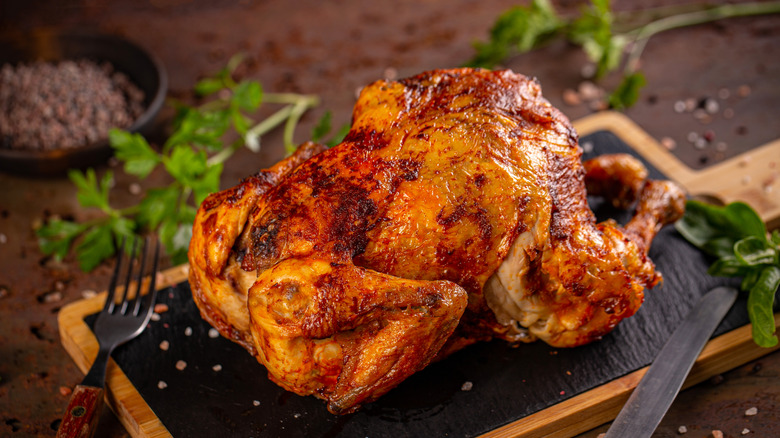 A rotisserie chicken sits on a slate cutting board with a knife and fork nearby