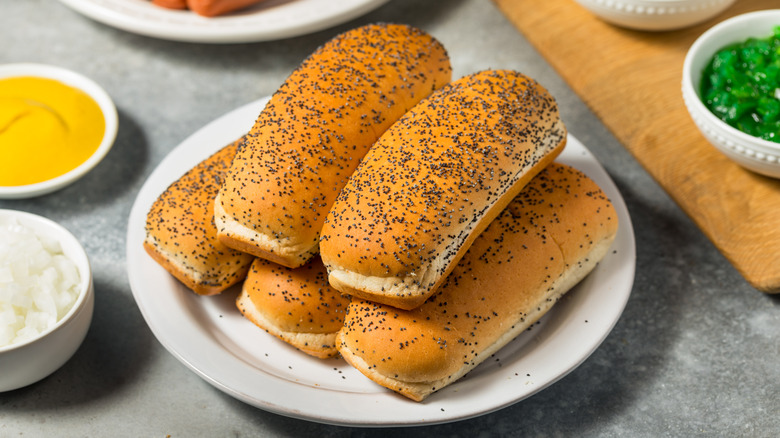 A plate of poppy seed hot dog buns rests next to hot dogs and various toppings