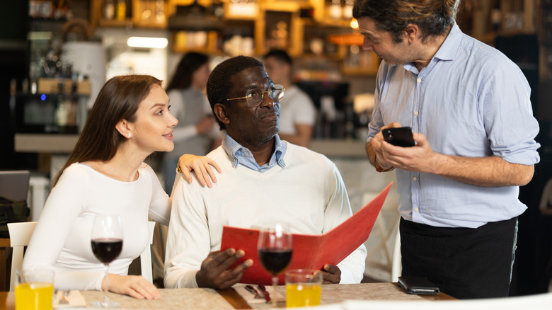 A waiter explains a menu item to two diners in a restaurant