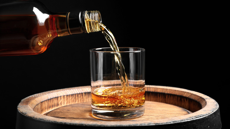 Bourbon pouring from bottle into glass sitting on a barrel
