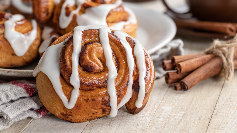 A cinnamon roll, drizzled with icing, sits on a wooden surface, in front of a white plate