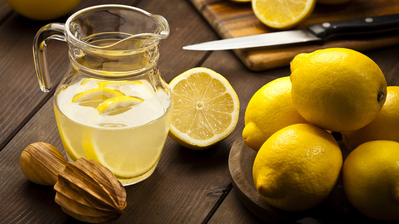 A pitcher of lemonade sits next to a juicer and a pile of lemons on a wooden table