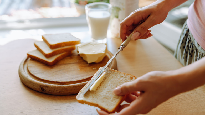 Someone butters a slice of bread, with other bread slices, butter, and a glass of milk in the background