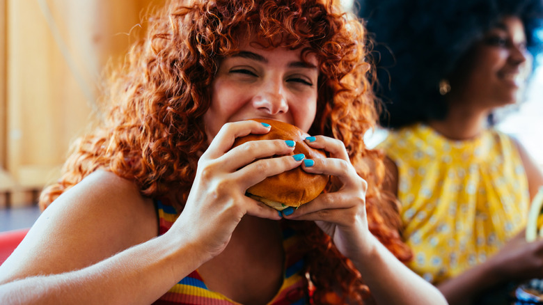 A diner with red curly hair takes a bite of a cheeseburger at a restaurant
