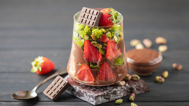 Viral Dubai chocolate and strawberry cup in glass, surrounded by chunks of chocolate, whole berries and pistachios
