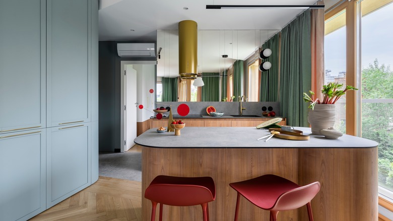 A midcentury modern kitchen with a rounded island, gold cylindrical range hood, and bright red chairs and decor pieces