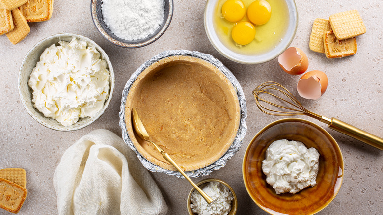 Cheesecake ingredients spread out on a counter, including eggs cracked into a bowl, softened cream cheese, flour, butter, and a cookie crumb crust
