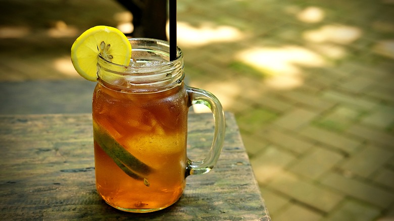 A clear glass mason jar mug with a handle holds iced tea and is garnished with a lemon wedge