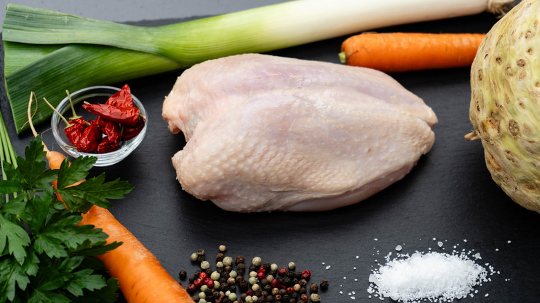 Full chicken breast surrounded by carrots, leeks, parsley, peppercorns, salt, and a small bowl of chiles on a black surface