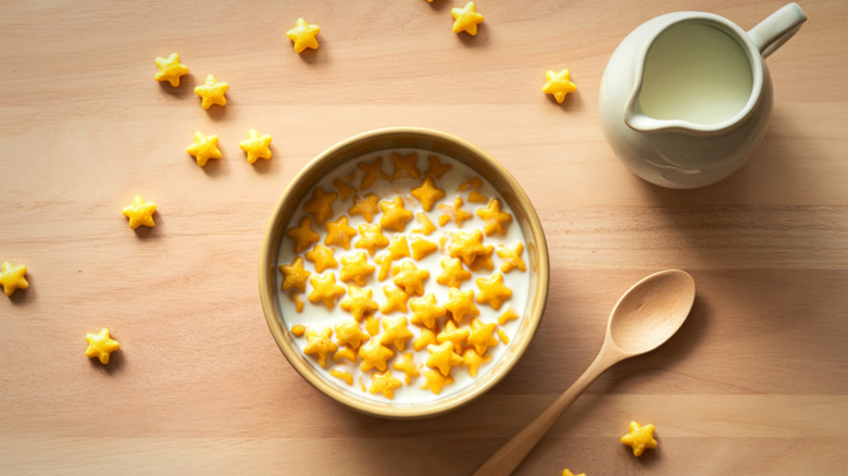 A bowl of star shaped cereal with milk, a wooden spoon, and scattered pieces of cereal surrounding it