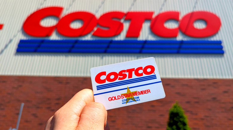 Person holding up a Costco membership card in front of the Costco sign