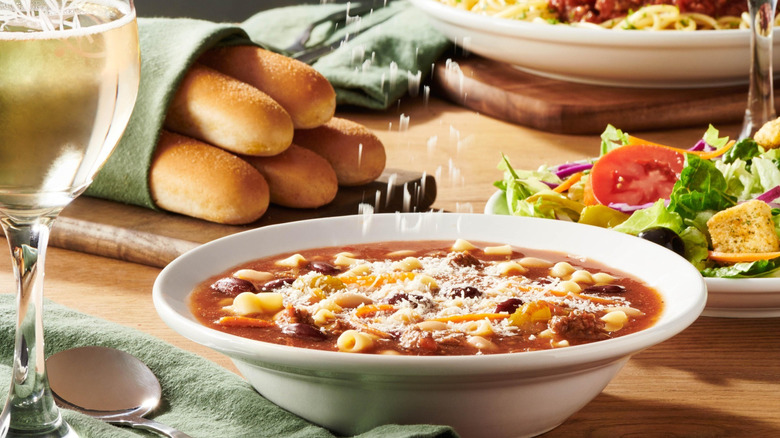 Someone shreds cheese into minestrone soup, next to a large salad and a basket of breadsticks