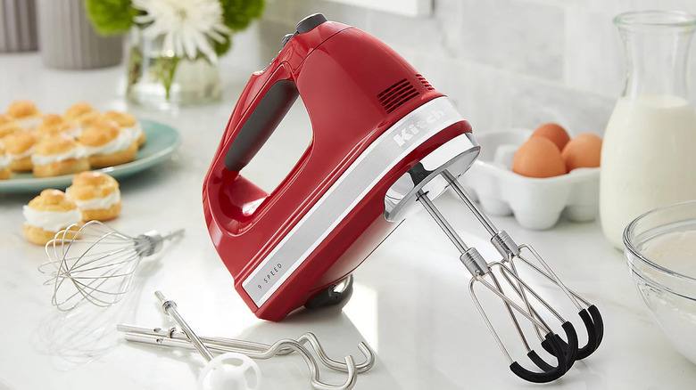 A KitchenAid hand mixer sits on a white countertop surrounded by attachments and baking items