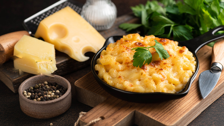 Single serving of mac and cheese in cast iron skillet next to wedge of cheese and a grater, a bowl of peppercorns, and herbs on a wooden board
