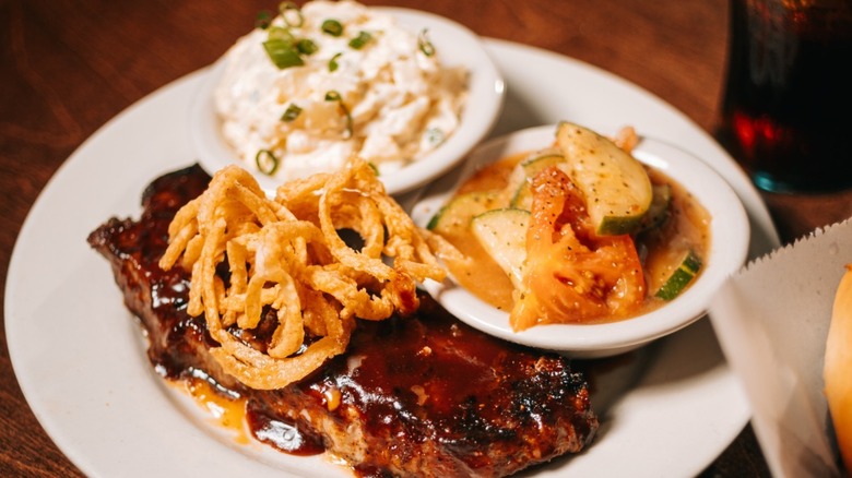 A mesquite barbecue glazed steak with fried onions, a side of sliced vegetables, and potato salad at Logan's