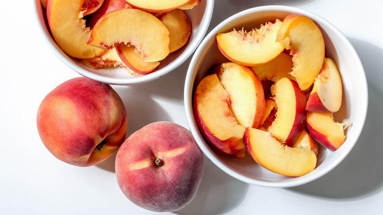 Two bowls contain sliced peaches while two whole peaches sit next to them