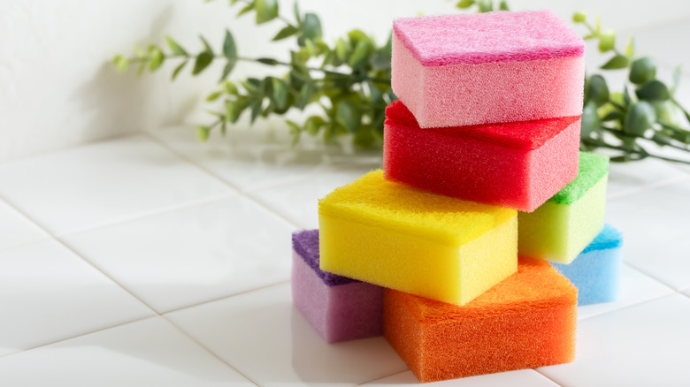 A stack of multicolored dish sponges sit on a counter with fake eucalyptus in the background
