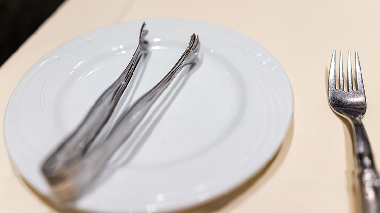Churrasco tongs on a white plate placed on a cream tablecloth next to a metal fork
