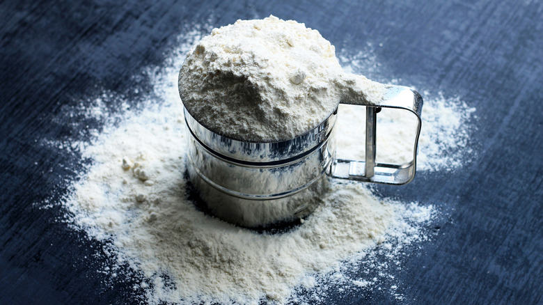 A mug-style flour sifter filled with flour sits atop a pile of flour on a blue surface