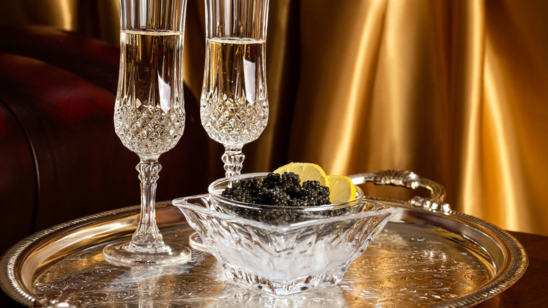 a small bowl of caviar sits next to lemon wedges and two glasses of wine in champagne flutes