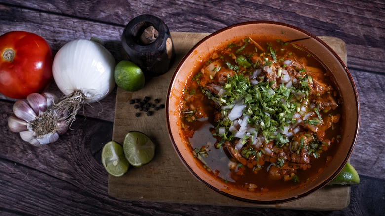Birria meat in a pot next to vegetables, aromatics, lime, and peppercorns.