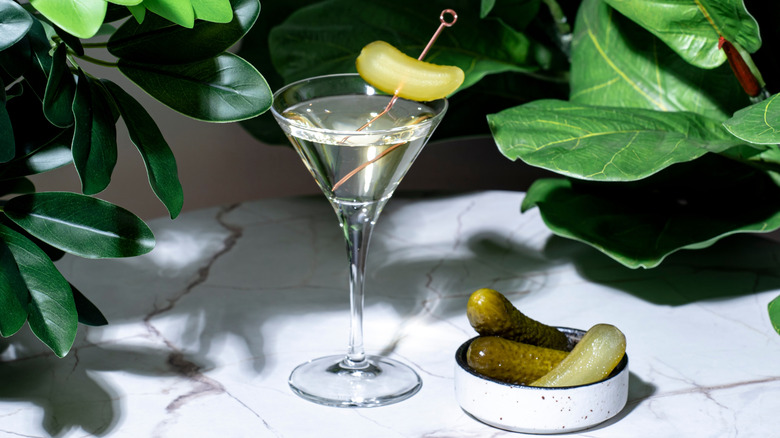 Pickle martini on a marble table next to a small bowl of pickles surrounded by leaves