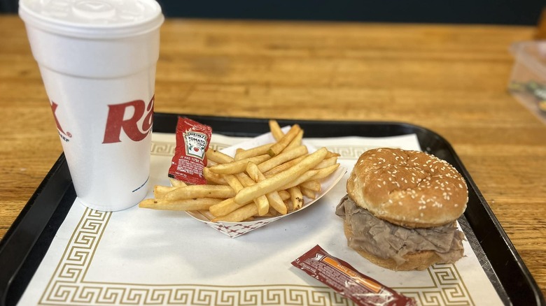 A roast beef sandwich, soda, french fries, and a packet of barbecue sauce from a Rax restaurant sit on a black tray