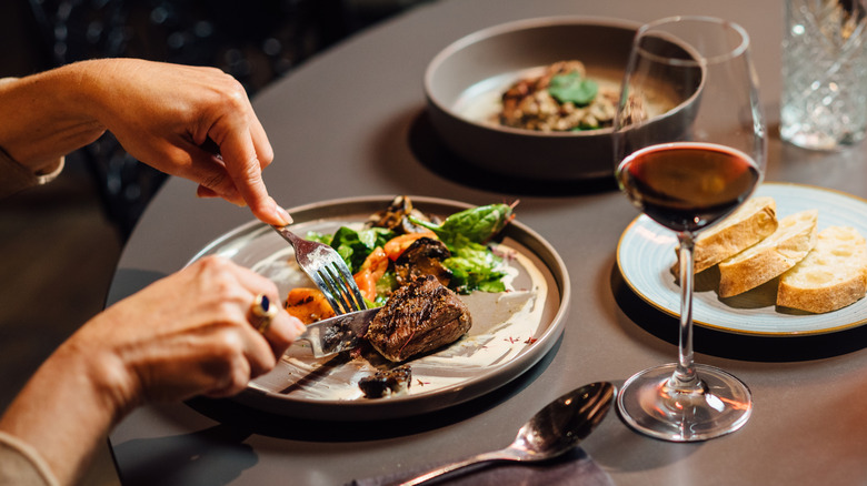 A diner at a find dining restaurant cuts into a steak on a plate next to a glass of red wine